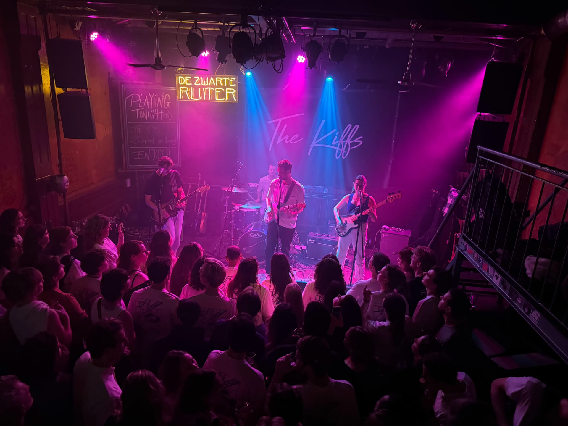 The Kiffs performing on stage in pink and blue stage light in front of a full crowd at De Zwarte Ruiter.